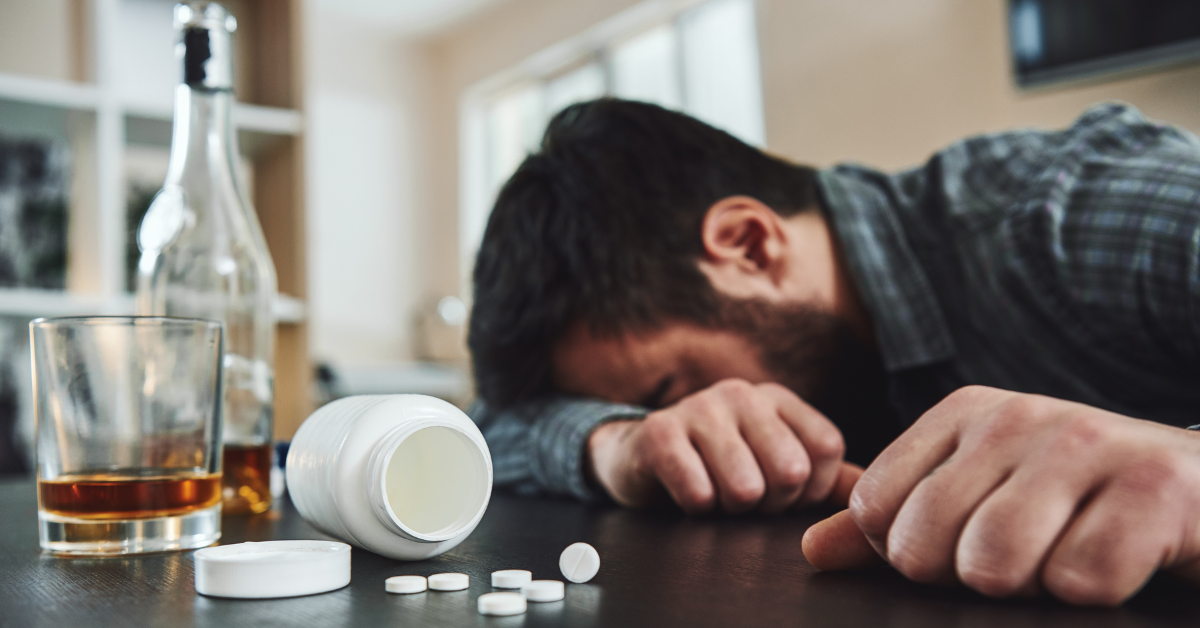 A man in denial is lying next to a bottle of alcohol and pills.