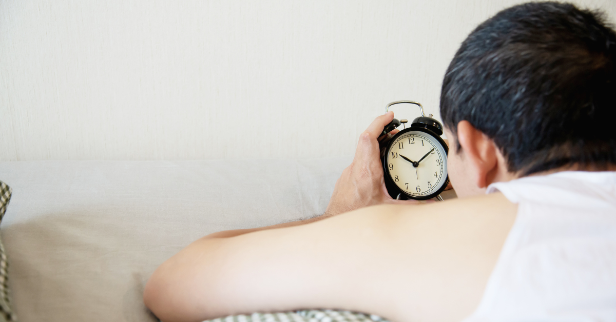 A patient man is laying in bed looking at an alarm clock.