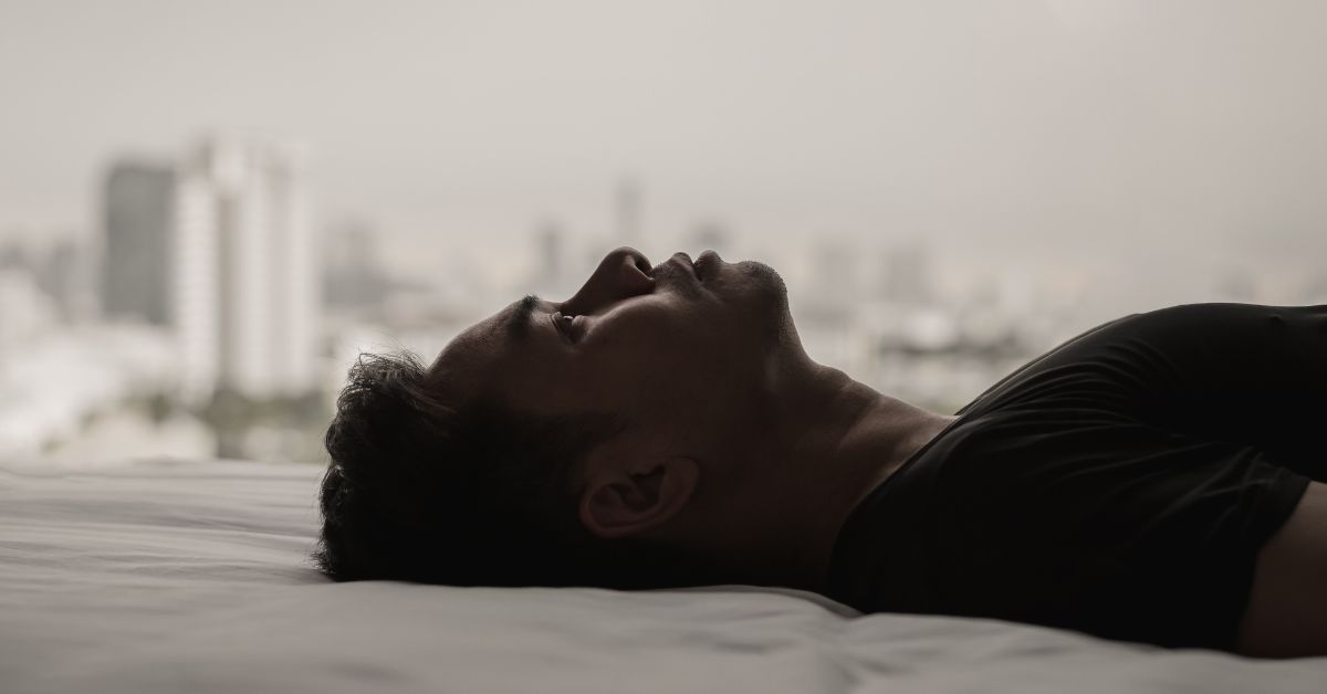 A man with a mental disorder, potentially influenced by alcohol abuse, is laying on a bed with a city in the background.