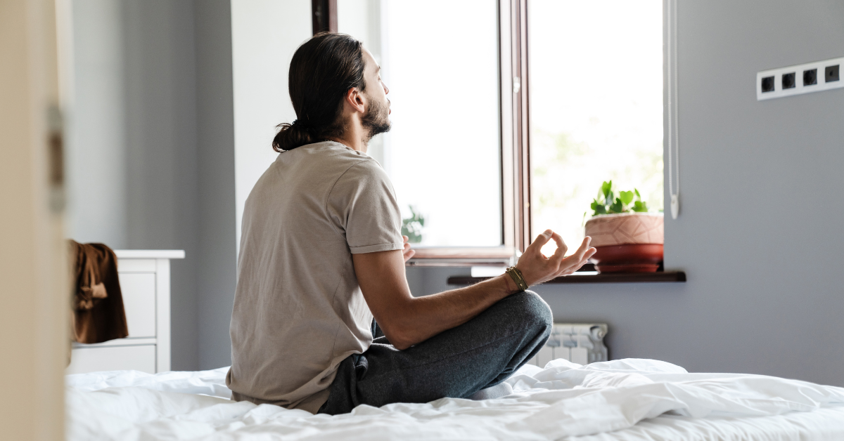 A man is practicing meditation, utilizing coping skills for addiction, on his bed in front of a window.