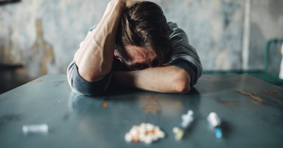 A man struggling with addiction sits solemnly on a table surrounded by pills, facing the harsh reality of tough love.