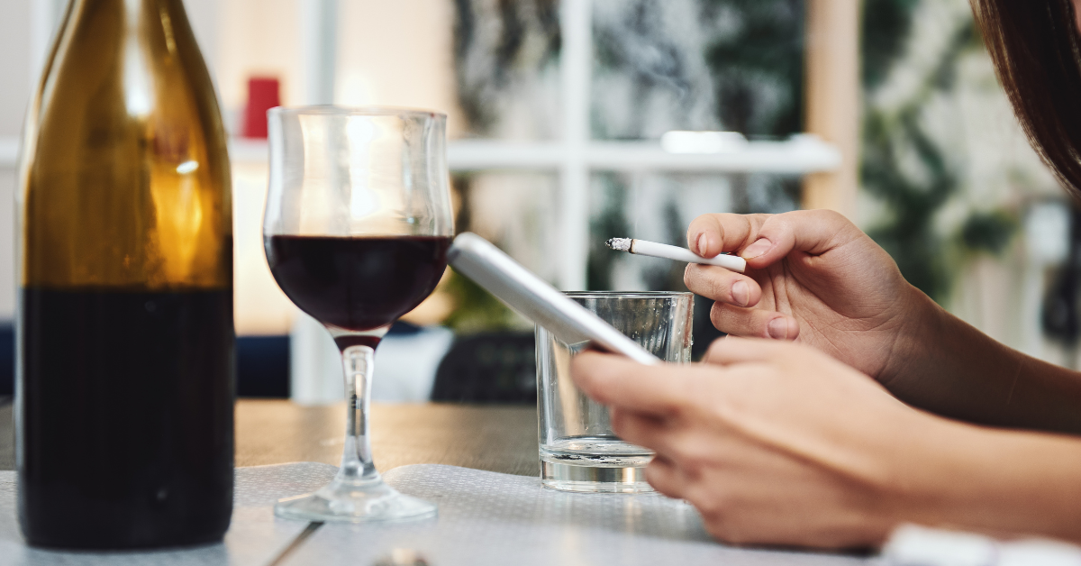 A woman looking on her phone how to overcome addictive personality. She is smoking a cigarette while holding a glass of wine.