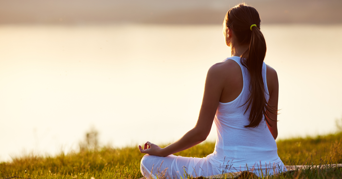 A woman is practicing selfcare in addiction recovery by meditating in the grass near a lake.