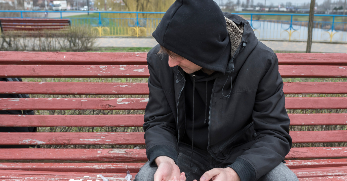 A man, seemingly lost in his addiction, wearing a hoodie and sitting on a bench.