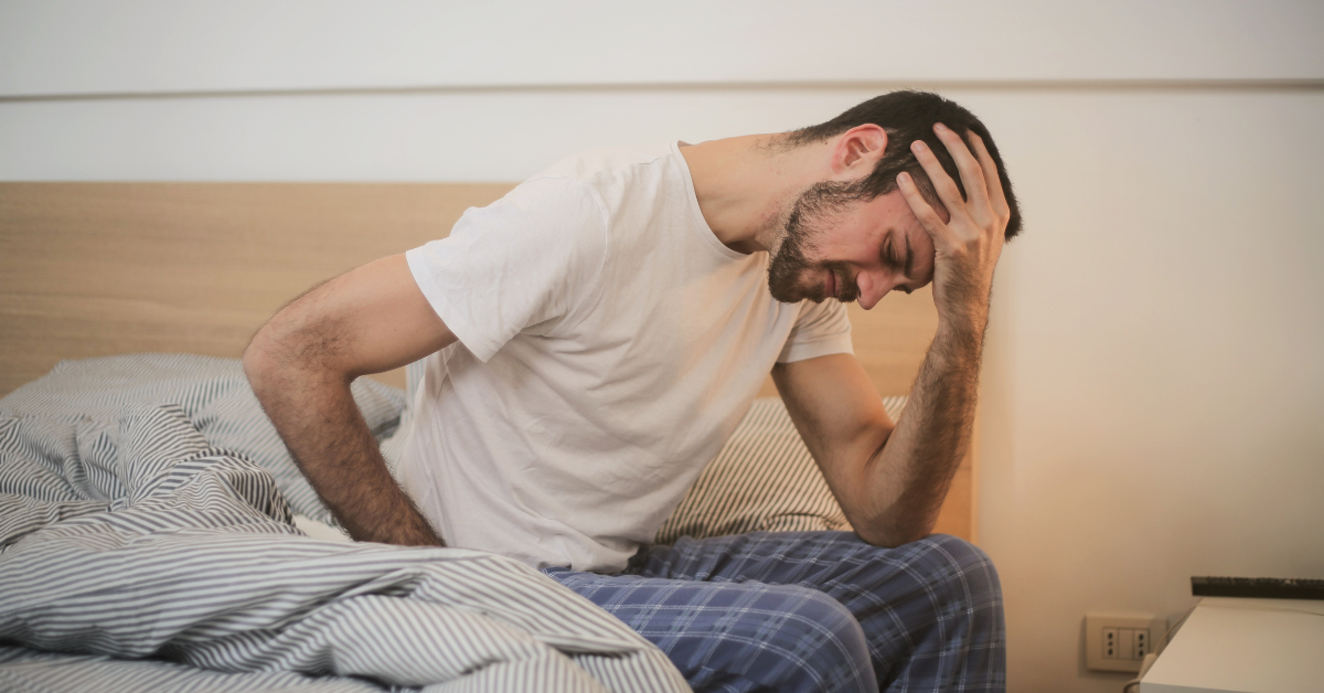 A man in pajamas is sitting in bed strugling with sobriety fatigue.