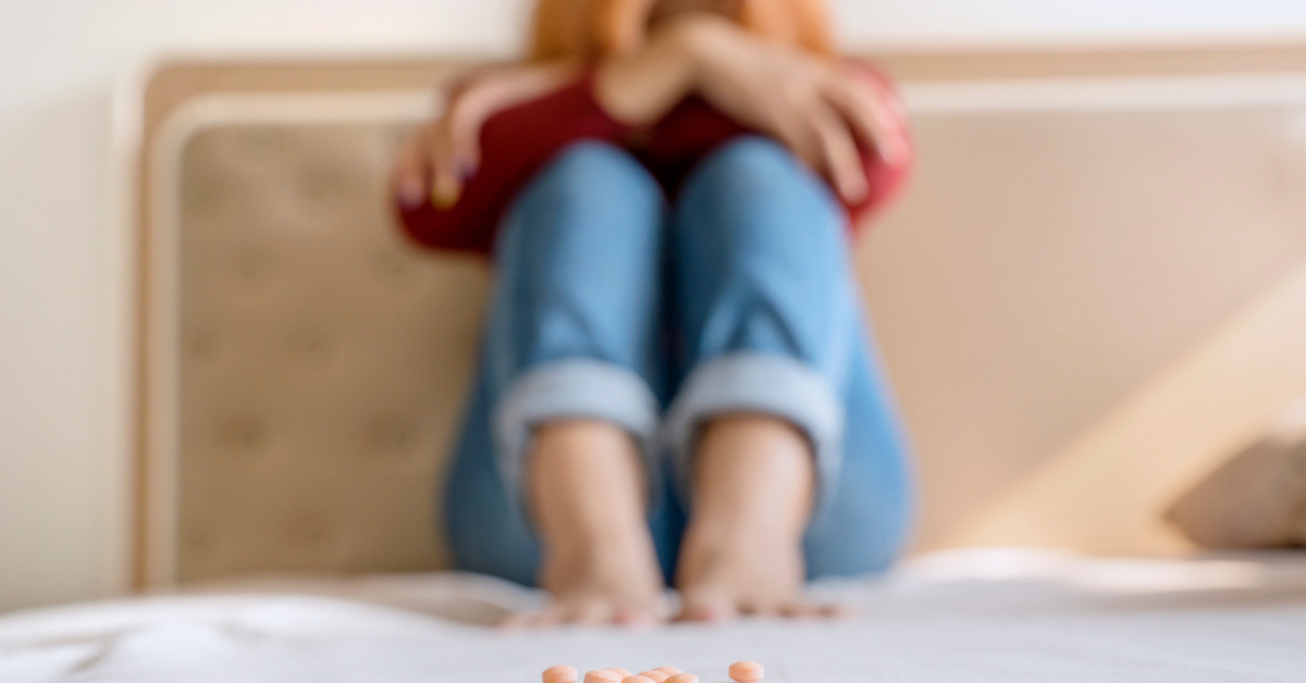 A teenager is sitting on a bed, surrounded by pills, illustrating the potential factors that influence teen drug use.