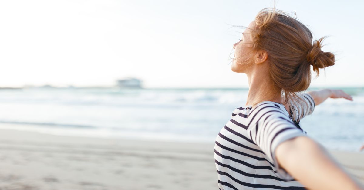Woman standing on the beach, experiencing freedom and true liberation in sobriety with arms outstretched.