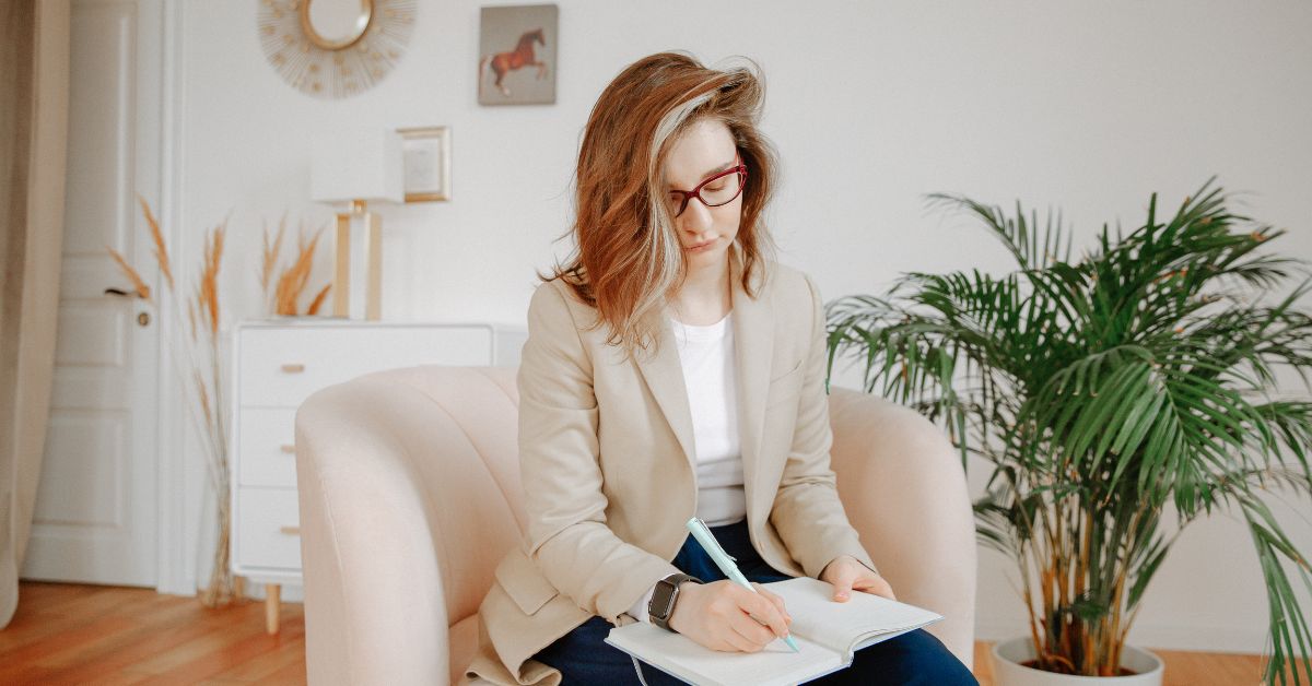 A mother is sitting in a chair and writing in a notebook, completely engrossed in her thoughts taking care of herself.