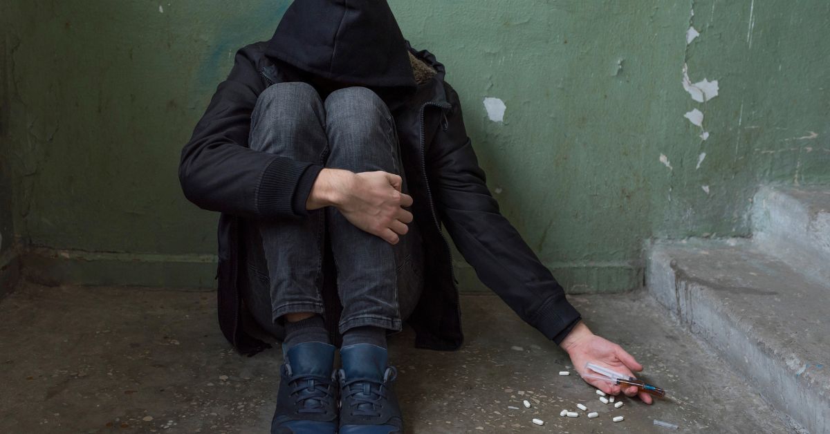 A teenager in a hoodie with signs of drug use sitting on the steps.