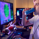 A teenager drinking from a bottle in front of two monitors.