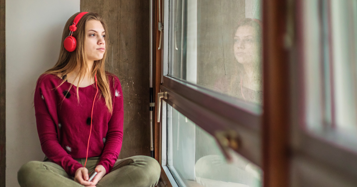 A teenage girl, lost in music with headphones, gazes out the window, feeling hopelessly alone. This solitude points to one of the four traits that increase the risk of addiction: isolation.