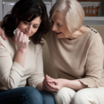 An older mother sits beside her grown daughter on a couch, one arm around her shoulders and holding her hand while the daughter wipes away tears.