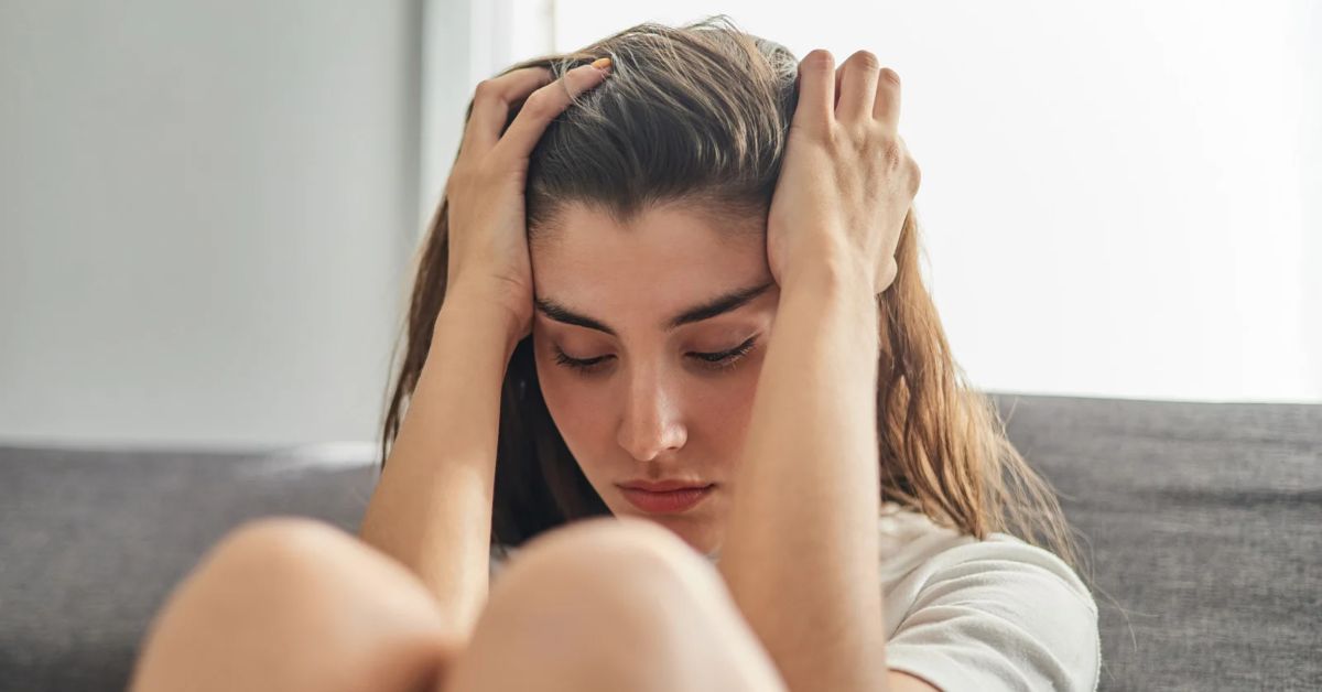 A woman seeking help overcoming addiction sits on a couch with her hands on her head. 