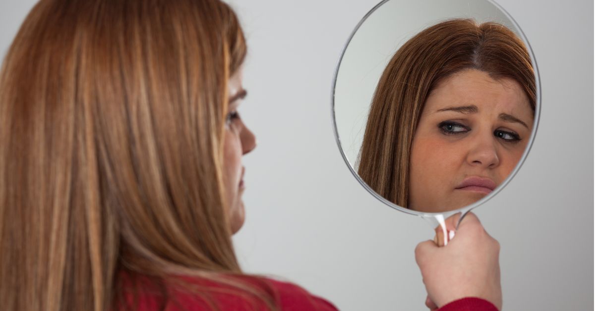 A woman reflecting on her appearance in a mirror, struggling with low self-esteem, contemplates the factors leading up to alcoholism.