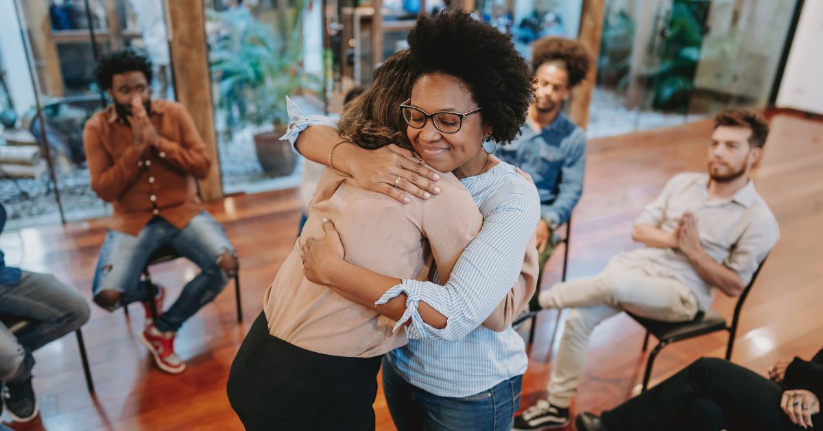 A woman is resiliently hugging another woman in a meeting room to cope with their recovery.