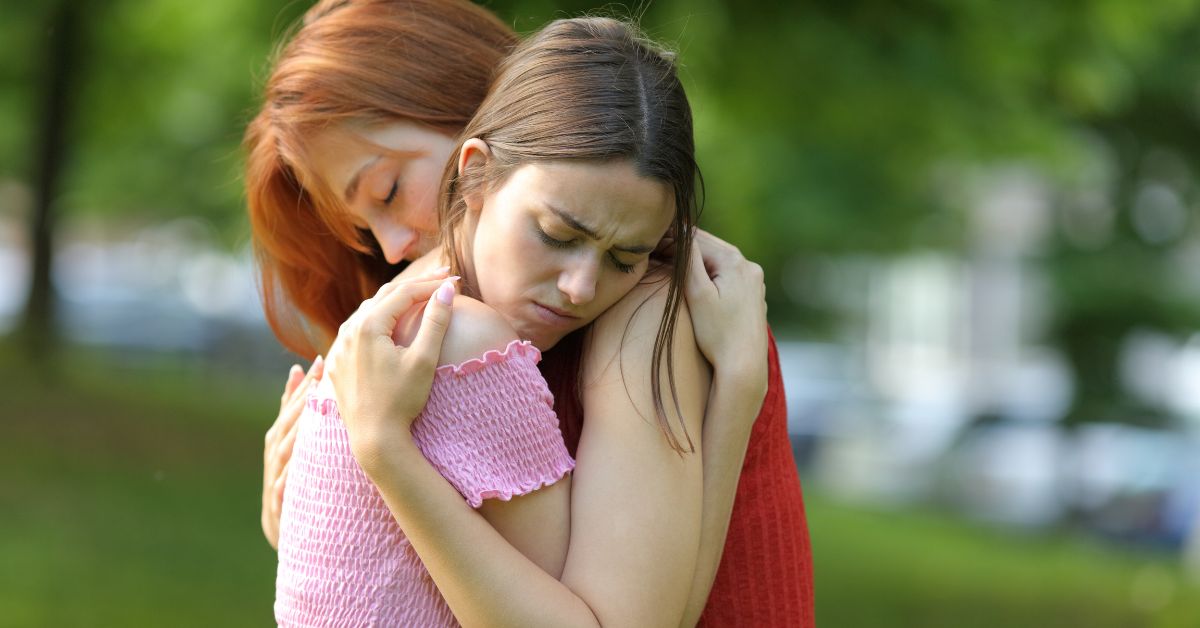 Two women hugging each other in a park, offering support to an alcoholic child.