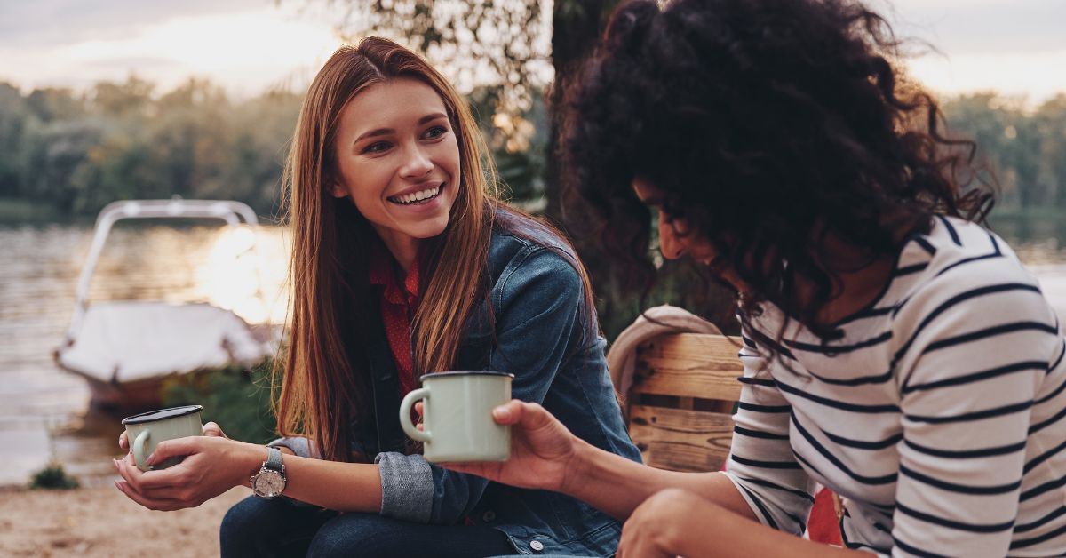 Two women sitting on a bench near a lake, enjoying coffee as part of their self-esteem boosting recovery journey.