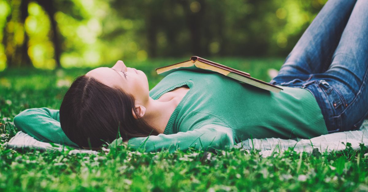 A woman managing her withdrawal symptoms in early recovery engrossed in her book, peacefully reclines on the grass.