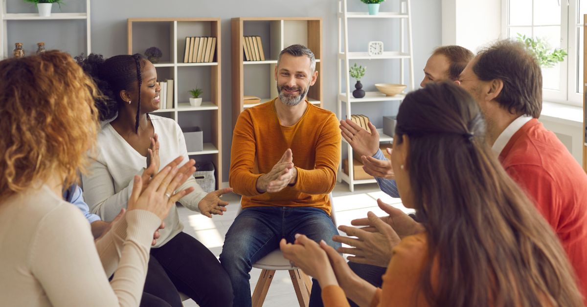 A group of emotionally unavailable people sitting around a table and clapping while attending a support group for alcoholics.