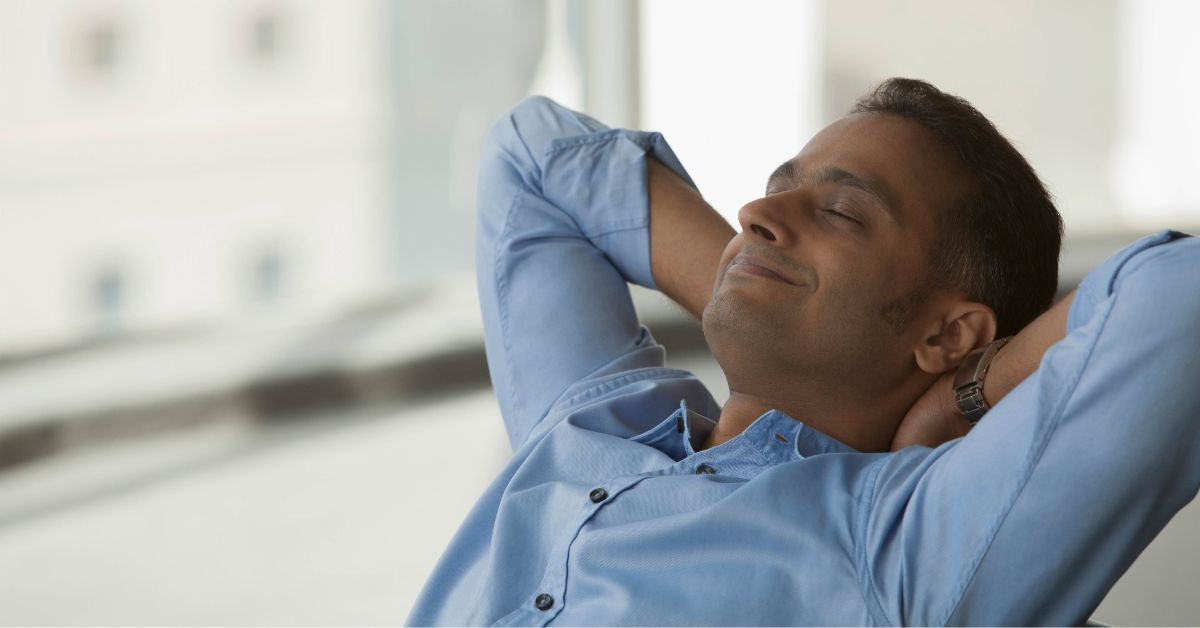 A man in a blue shirt embracing sobriety relaxes with his hands on his head.