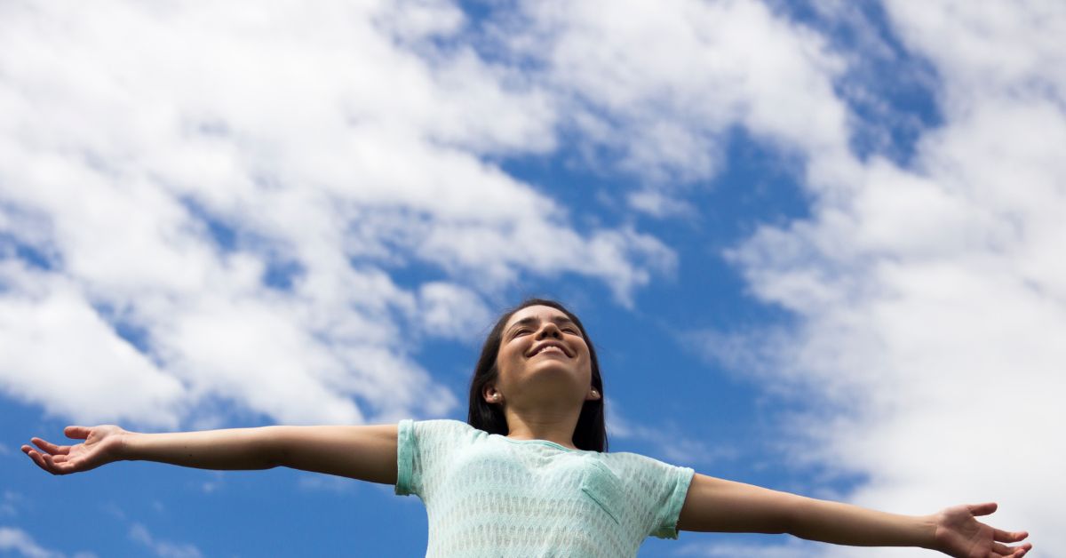 A woman overcoming shame with her arms outstretched against a blue sky.