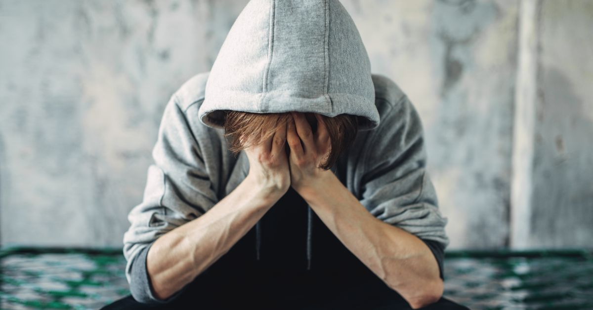 A man in a hoodie is covering his face with his hands, displaying withdrawal symptoms after coming down on crack. 