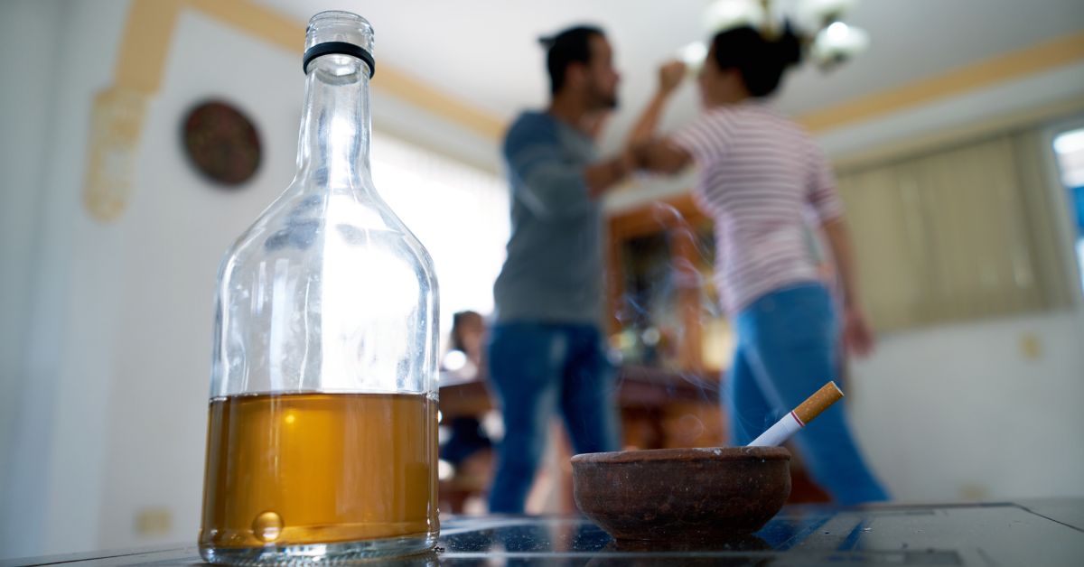 A bottle of alcohol and a cigarette on a table, symbolizing why an alcoholic cannot love with two people in the backgrounding fighting