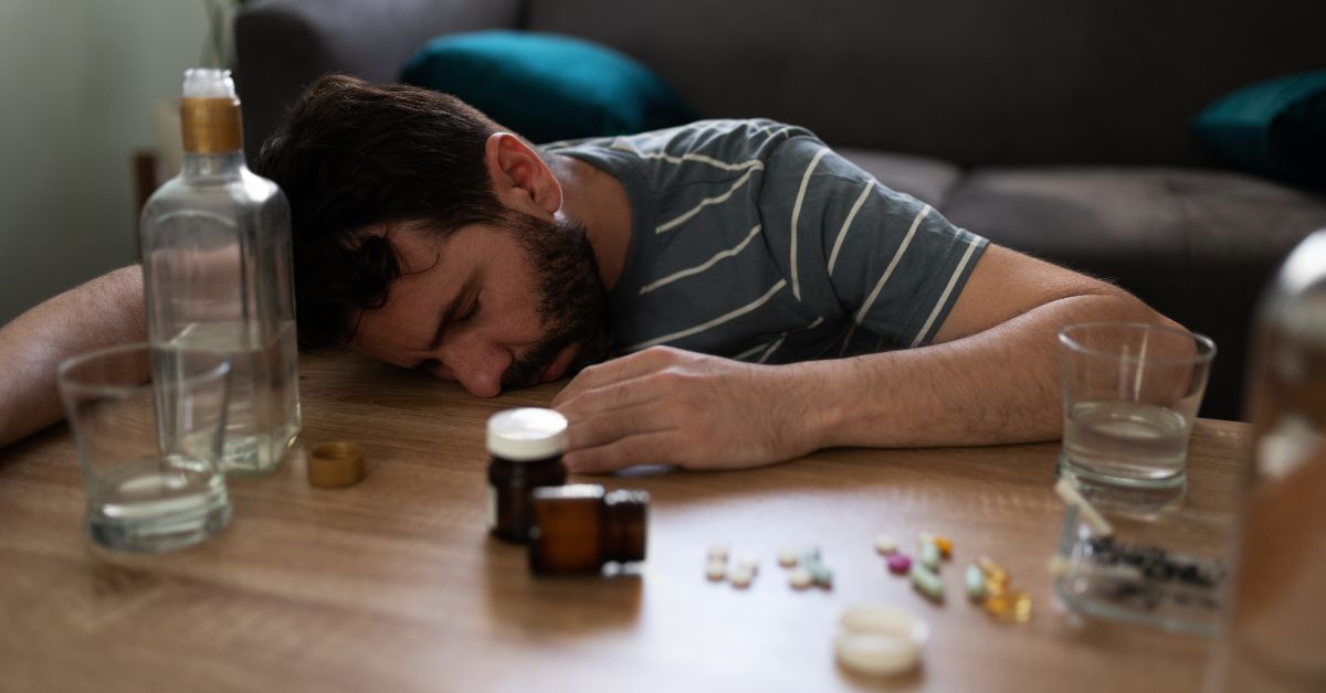 A man is engaging in unhealthy coping skills by lying on a table with pills and alcohol.