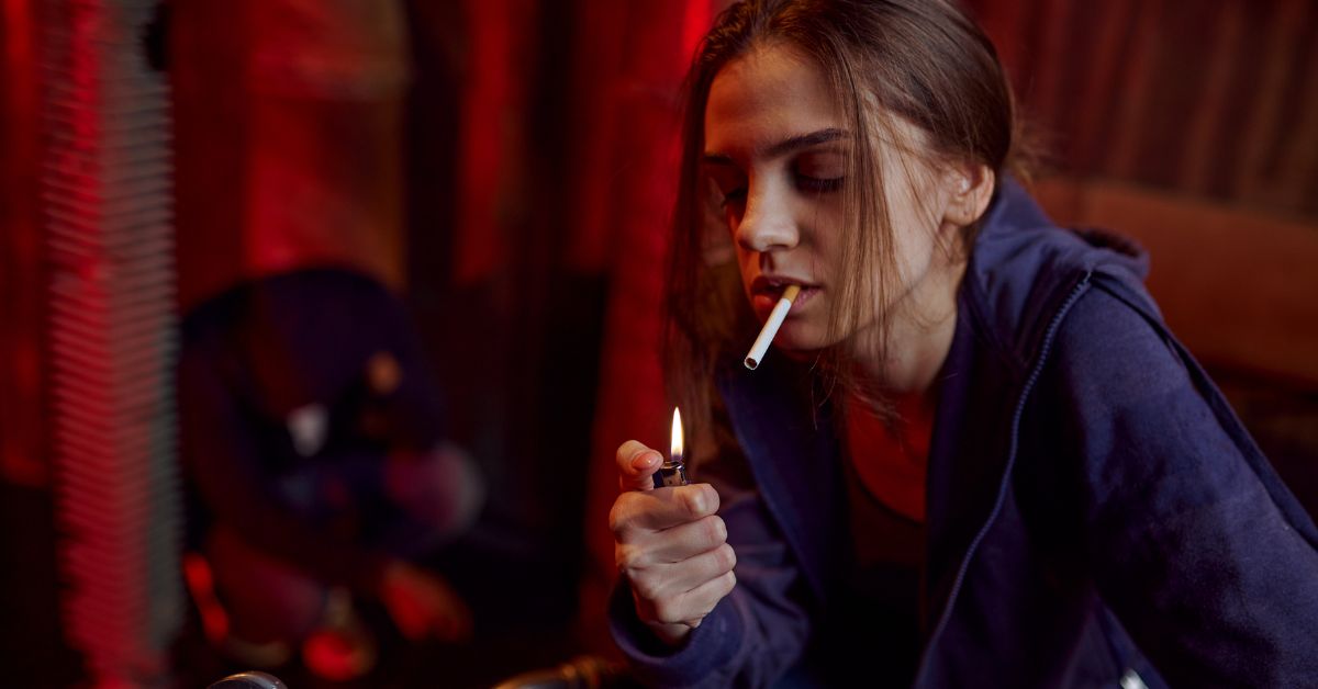 A girl is smoking a cigarette in a dimly lit room, exhibiting warning signs that her drug rehabilitation might not be successful.